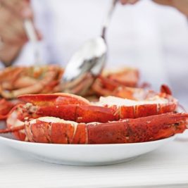 A person in a white shirt is eating a seafood meal consisting of a large red lobster on a white plate. The individual's hands are cutting into the food using a fork and spoon. The table is white and has a spoon resting on it.