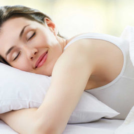 A woman with light brown hair is peacefully sleeping on her side with a white pillow and white bedding. She is wearing a white tank top and smiling gently with a relaxed expression on her face, showcasing how to get the best night's sleep. The background is softly blurred, creating a serene atmosphere.