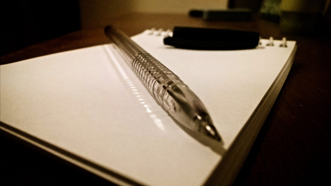 Close-up of a clear plastic pen resting on a blank spiral notebook on a wooden table. The notebook is open, and a black wristwatch is placed beside it, perhaps awaiting notes on how to get the best night's sleep. The image has a warm, dim-lit ambiance, with the focus on the pen.