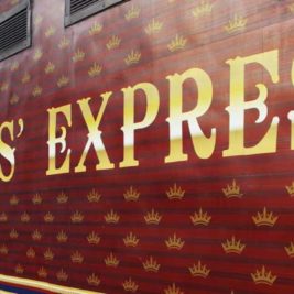 A person in a hat and floral dress stands next to the Maharajas' Express train, which is decorated with a maroon and gold crown pattern. The train's name is prominently displayed in large, ornate yellow letters. The scene evokes luxury and elegance.