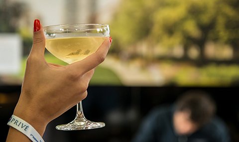 A person's hand with red nail polish is holding a champagne glass filled with a light-colored liquid. The person, gearing up for Salon Privé, is wearing a white wristband. The blurred background shows an outdoor scene with greenery and a person in a dark-colored shirt.