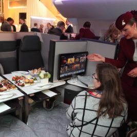 A flight attendant in a maroon uniform assists a seated passenger in business class, demonstrating the features of an in-flight entertainment system. The long haul luxury setup includes plated meals on a fold-out table, with other passengers and modular seating visible in the background.