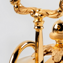 Close-up of an ornate, gold-colored object featuring intricate detailing and a luxurious finish. The image showcases a curved handle and decorative elements with a glossy appearance, suggesting high craftsmanship.