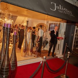 A fancy inauguration event at Julien's Interiors. The entrance features a red carpet, velvet rope barriers, large decorative vases, and a chandelier inside the store. People are mingling inside while a man stands at a podium outside the door, speaking into a microphone.