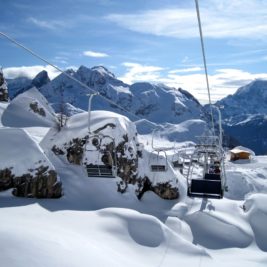 A picturesque snowy mountain landscape featuring a ski lift with several empty chairs suspended over the snow-covered terrain. In the background, tall, rugged mountains rise against a clear blue sky with wisps of clouds.