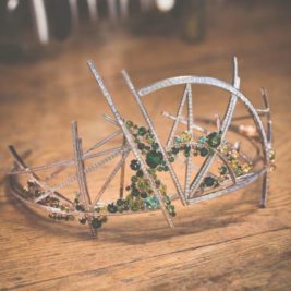 A delicate tiara with an abstract, angular silver framework adorned with green and clear gemstones sits on a wooden surface. The design is modern and artistic, featuring intersecting lines and scattered gems.