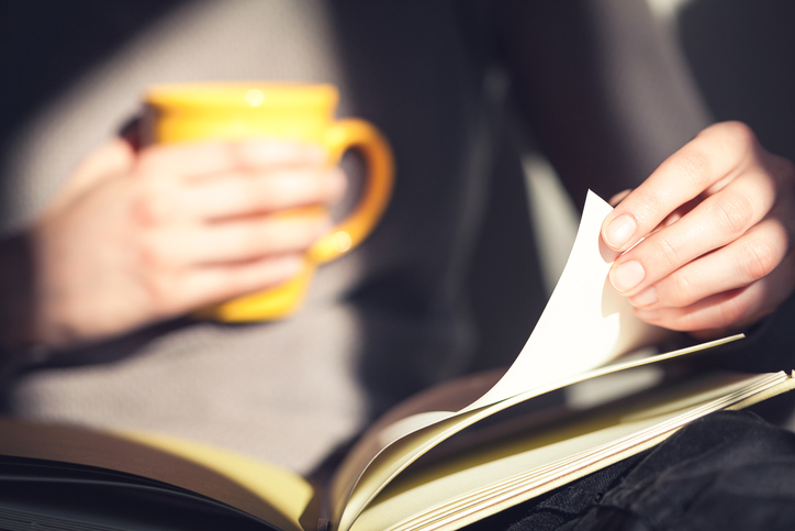 A person holds a yellow mug in one hand and turns the page of a book with the other. The warmly lit scene creates a cozy and relaxed atmosphere, exemplifying how to be the perfect host.