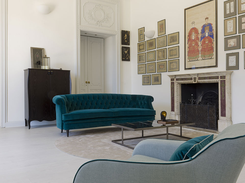 A modern living room with a teal tufted sofa, a gray armchair, and a glass coffee table on a circular beige rug. The space boasts bespoke furniture, including a black cabinet and decorative fireplace adorned with framed artwork on the walls. The room features white walls and a minimalist aesthetic.