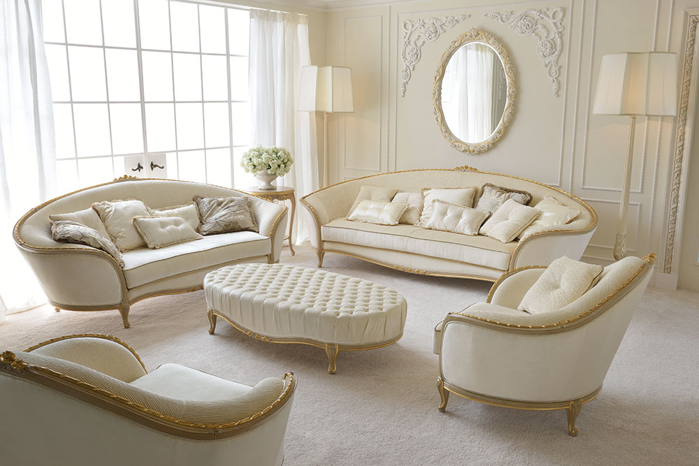 A luxurious living room with bespoke, cream-colored furniture, including two sofas, an armchair, and a tufted ottoman. The pieces feature gold accents. The room boasts large windows with white curtains, a mirrored frame on the wall, and elegant wall decor.