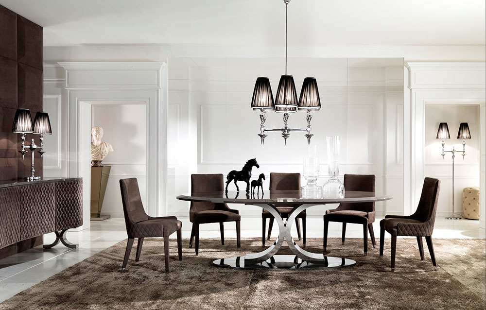 A modern dining room with a glass-top table and six bespoke brown upholstered chairs on a large beige rug. A black chandelier with multiple lampshades hangs overhead. Decorative horse statues are on the table, and a bust is displayed in the background. Walls are white.