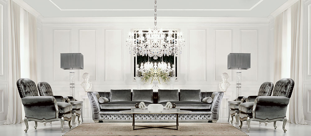 A luxurious living room features a large gray velvet sofa with matching bespoke armchairs. Two crystal lamps stand on either side of the sofa, while a grand chandelier hangs from the ceiling, illuminating the space. The room is decorated in white tones with a minimalist style.