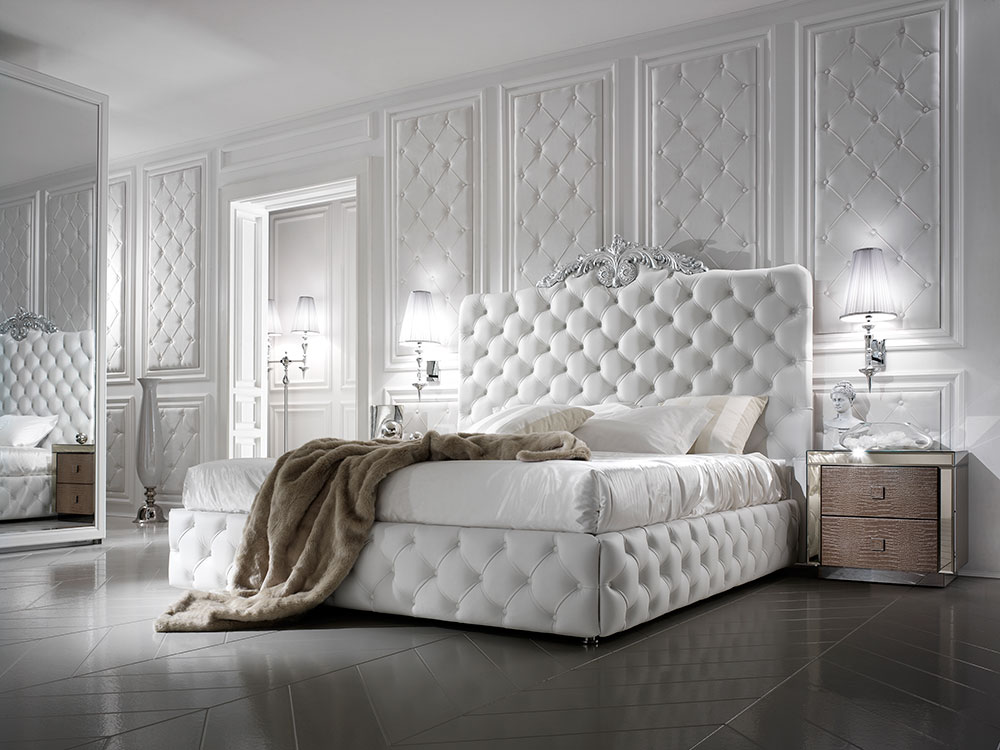 Luxurious white bedroom featuring a tufted upholstered bed with a fluffy blanket. The room has paneled walls, mirrored closet doors, modern bedside tables with lamps, and elegant decor from luxury Italian furniture designers, creating a cozy and opulent ambiance.