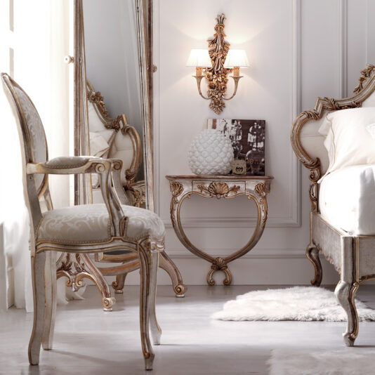 Ornate Rococo Wall Mounted Bedside Table A luxurious bedroom features antique, white and gold furniture. A large mirror stands next to a decorative chair with ornate carvings. The Ornate Rococo Wall Mounted Bedside Table holds a white textured vase and a book. The room is softly lit by a wall sconce above the table, and a plush rug lies on the floor.