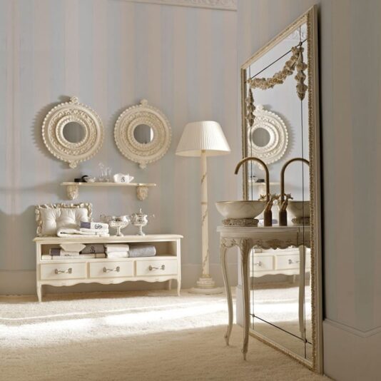 An elegant, vintage-style bathroom featuring a large framed mirror, a vanity with two ornate brass faucets, a freestanding sink, an Italian 3 Drawer Cabinet for extra storage, a decorative bench with storage, a floor lamp, and two round decorative wall mirrors. The room is adorned in a soft, neutral color palette.