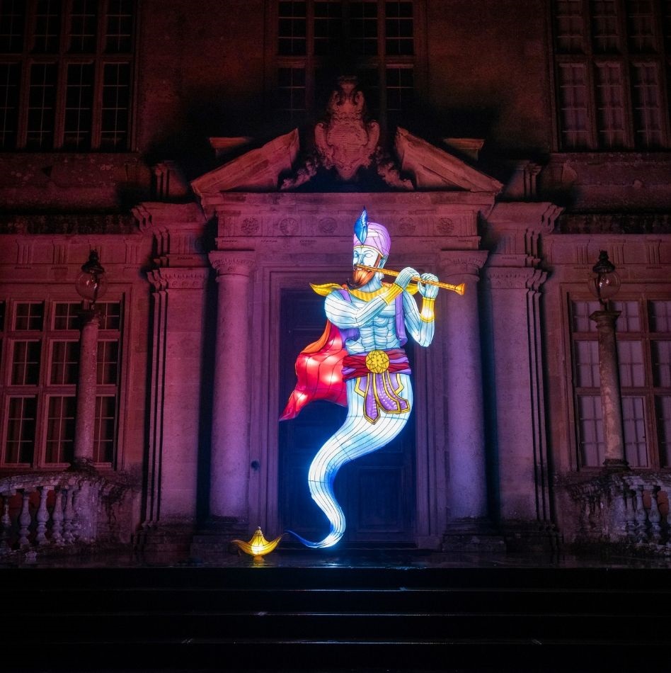 A vibrant and illuminated sculpture of a genie rising from a golden lamp is displayed in front of an ornate and dimly lit building. The genie has a blue body, a red sash, and a golden turban, holding a flute, creating a magical atmosphere.