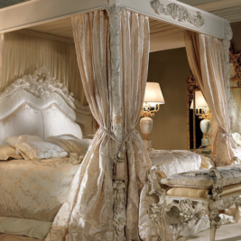 Luxurious bedroom with a canopy bed adorned with ornate carved details and draped in elegant curtains. The bed, an ex display piece, is dressed in opulent bedding, and a matching bench sits at its foot. Two ornate nightstands with lamps flank the bed, and a large decorative mirror stands in the corner.