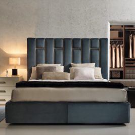 A modern bedroom, spotlighted in the Top 10 Interiors blog, features a large bed with a tall, upholstered headboard in dark blue. The bed is adorned with multiple pillows and neutral-colored bedding. A bedside table with a lamp is on the left, and an open closet with hanging clothes is in the background.