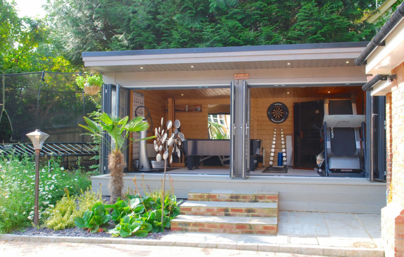 A modern shed with large glass doors open, revealing an interior equipped with gym equipment, a dartboard, and a console table. The exterior boasts a small garden with plants and steps leading up to this outdoor retreat. Trees and greenery surround the area.