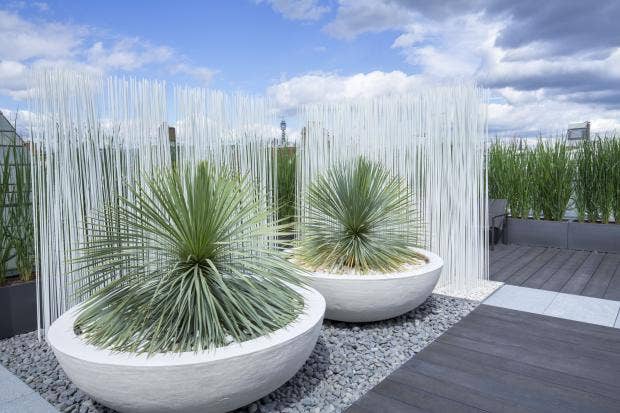 A modern garden features two large, round white planters with spiky green plants on a bed of gray gravel. Behind them is a decorative section of tall, thin white rods, and the garden is surrounded by greenery. A wooden deck is visible on the right side, leading to a cozy balcony area.