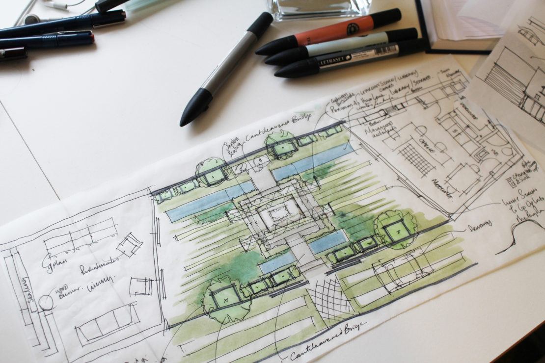 An architectural landscape design blueprint laid out on a desk with various pens, markers, and a notebook nearby. The sketch includes hand-drawn details and notes, showing pathways, greenery, and structural elements of an outdoor retreat, all accompanied by color highlights.