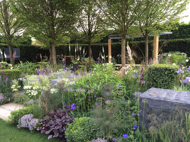 A lush garden scene serves as an idyllic outdoor retreat with tall trees providing shade and a variety of flowering plants in full bloom. The garden features a mix of colorful flowers, green shrubs, and stone elements, while a tall green hedge in the background frames the vibrant floral display.