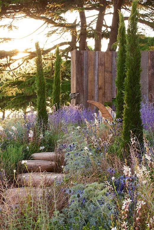 A rustic outdoor retreat features a path made from log steps surrounded by lush greenery, purple and white flowers, and tall, narrow trees. In the background, a wooden gate leads to a sun-dappled area where sunlight filters through the tall trees.