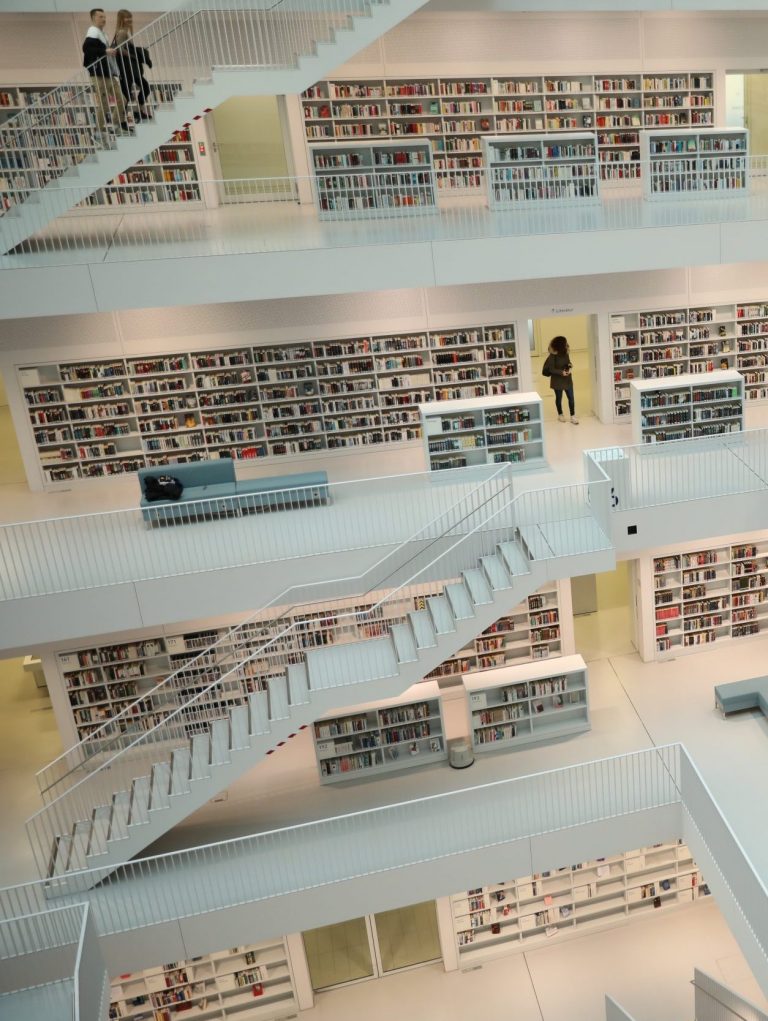 A multi-level modern library with white interiors and an open central space features cozy book nooks. Several floors of book shelves are visible, with staircases connecting the levels. A few people stand or walk, and the library is brightly lit.