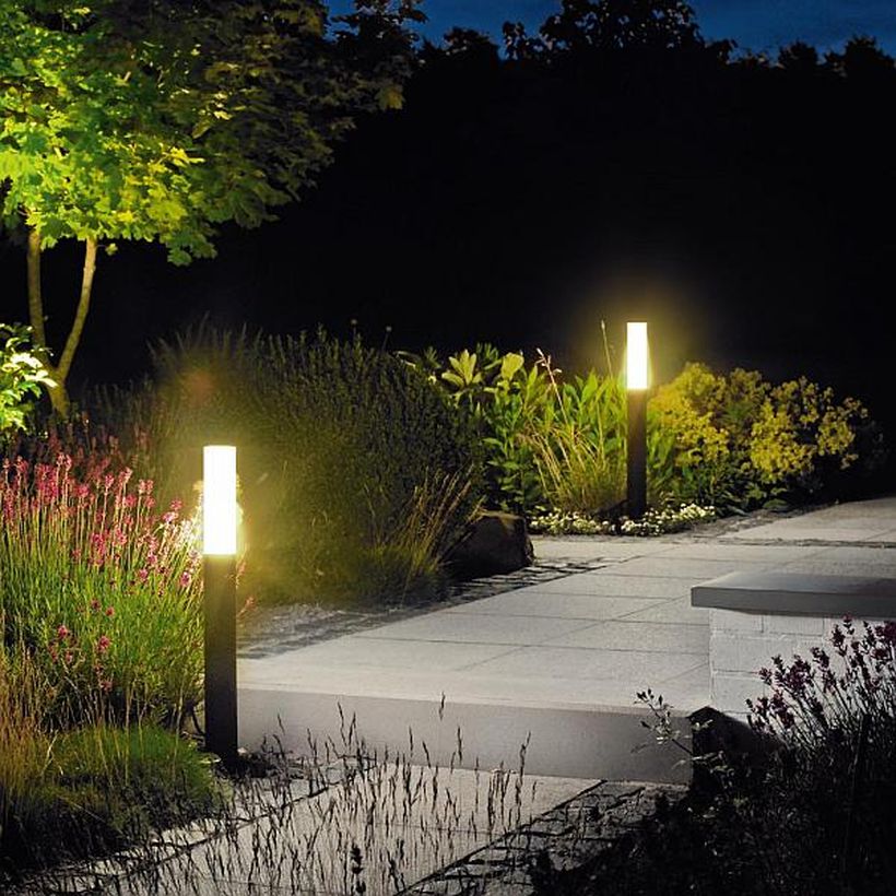 A nighttime garden scene with a lit pathway. The pathway is bordered by various plants and two modern, square pillar lights emitting a warm glow, illuminating the surroundings. With thoughtful outdoor lighting, a tree is visible in the background, partially lit by one of the pathway lights.