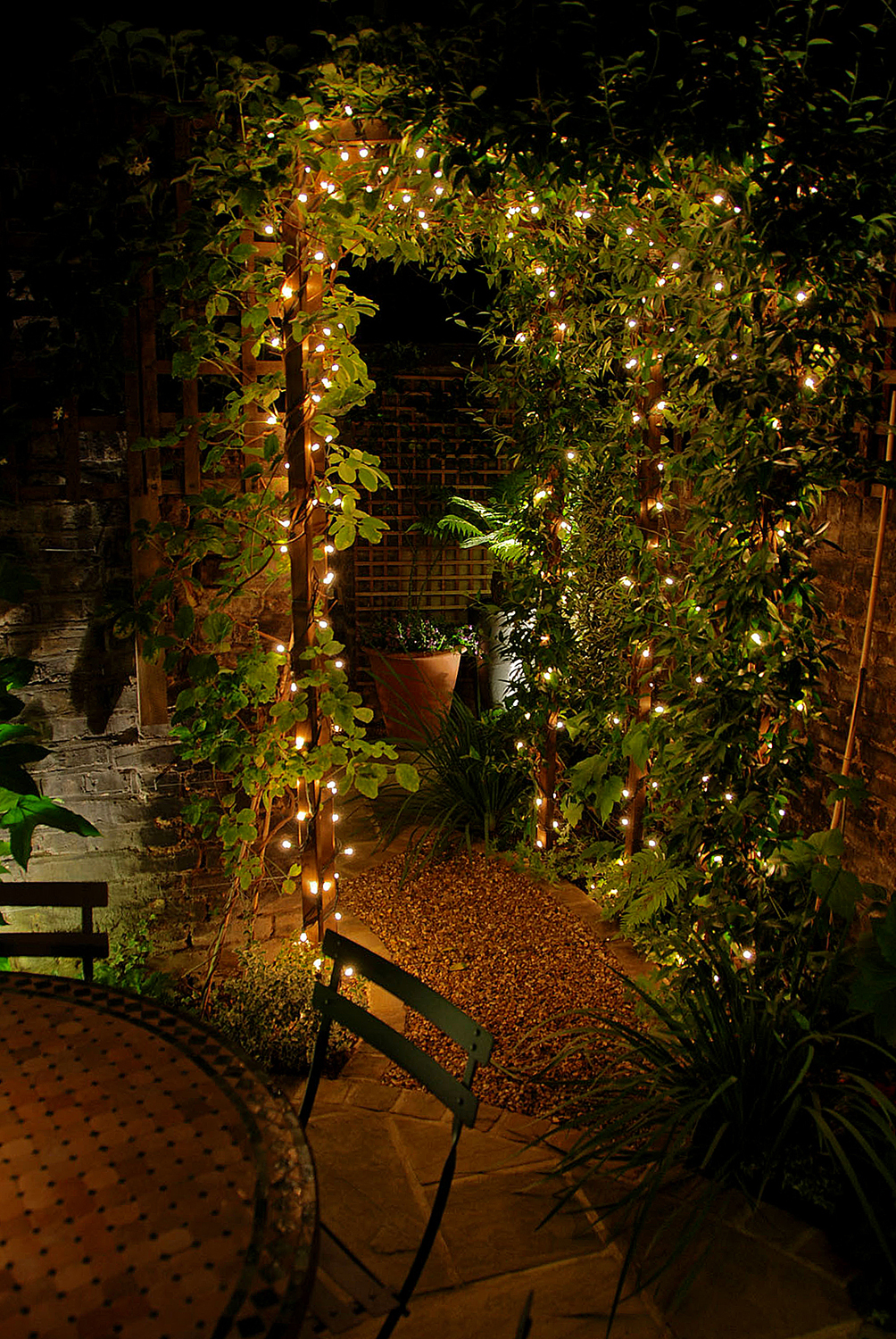 An outdoor garden scene at night, featuring an archway covered in lush greenery and adorned with strings of warm white fairy lights. The ground is covered with gravel and plants, and a round table with a chair is in the foreground, illuminated by soft outdoor lighting.