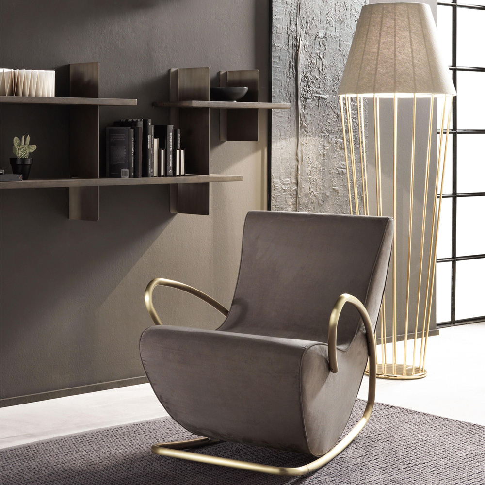 A modern, minimalist book nook featuring a grey rocking chair with gold armrests and a sleek geometric shape. Behind it, a textured grey wall with floating shelves displaying books and decor items. To the right, a tall floor lamp with a fabric shade and a gold frame.