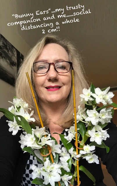 A woman with blonde hair and glasses smiles at the camera, holding two sticks with white flowers labeled “Bunny Ears.” Above them, text humorously comments on 2 cm social distancing. Dressed in a black and white outfit, she stands against neutral walls, hinting she's got "wishlist" items ready for any occasion.