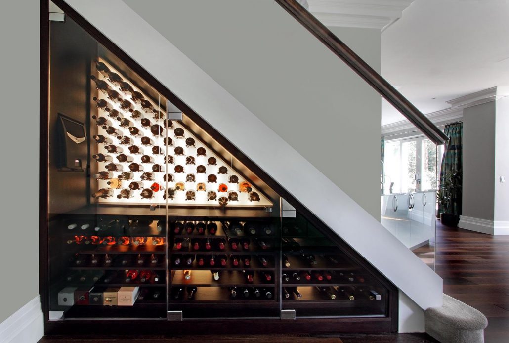 A modern home wine cellar with glass doors, built under the stairs. The cellar has multiple shelves filled with neatly arranged wine bottles, both vertically and horizontally. A portion of the living area with wooden floors is visible in the background.