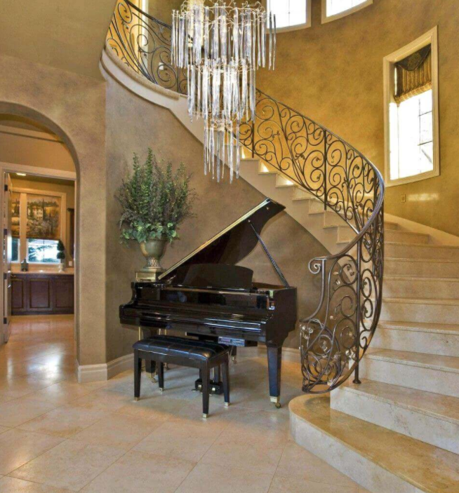 A grand piano with a matching bench is placed under the stairs at the base of an elegant spiral staircase with ornate wrought iron railings. A large chandelier hangs overhead, and a large potted plant sits nearby in a spacious, warmly lit interior with tan walls and tiled floors.