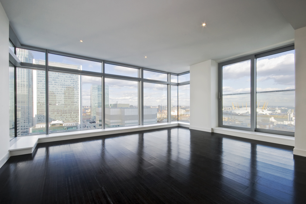 A spacious, modern living room with floor-to-ceiling windows offering panoramic views of a cityscape. The room features dark hardwood floors, white walls, and recessed lighting in the ceiling. Through an online consultation, you can explore virtual design options that enhance skyscrapers and the partly cloudy sky visible through the windows.