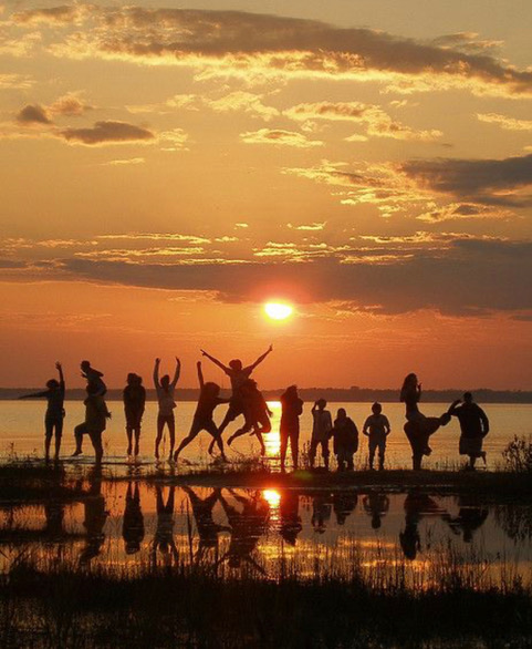 A group of people is silhouetted against a vibrant sunset sky, enjoying themselves by jumping and striking playful poses on the shore of a calm body of water. The sun is low in the sky, casting a warm orange glow and reflecting on the water's surface—it's the kind of moment that belongs on your wishlist.