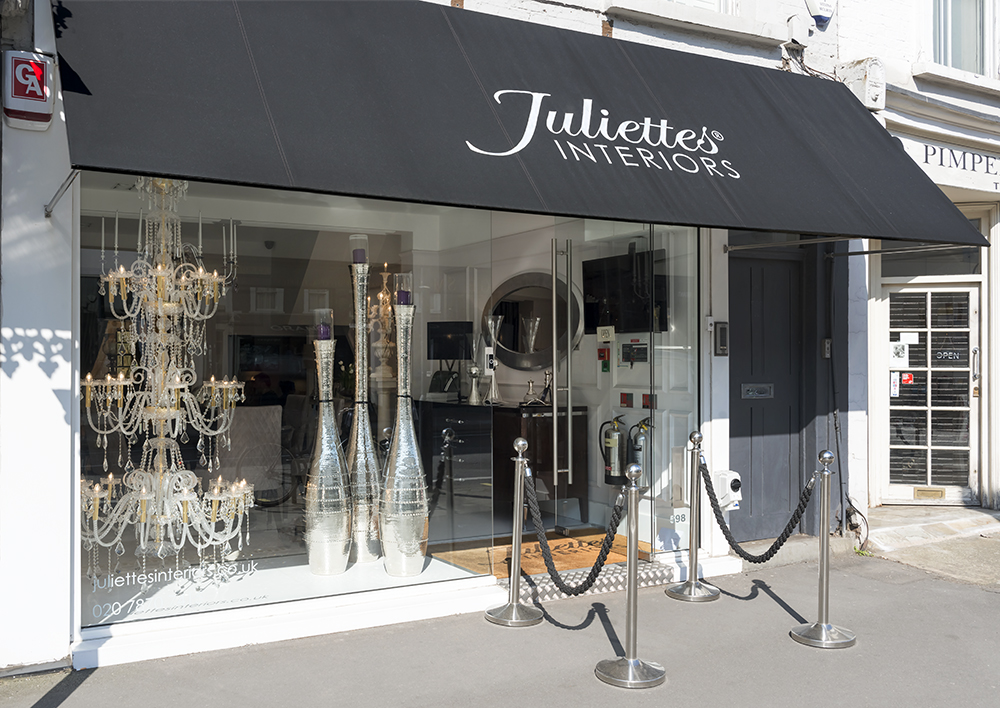 Storefront of Juliette's Interiors featuring a black awning with white logo and text. The window displays ornate crystal chandeliers, tall silver decorative pieces, and elegant furniture. The entrance has black stanchions with velvet ropes. Experience our expertise through online consultation for your interior design needs.