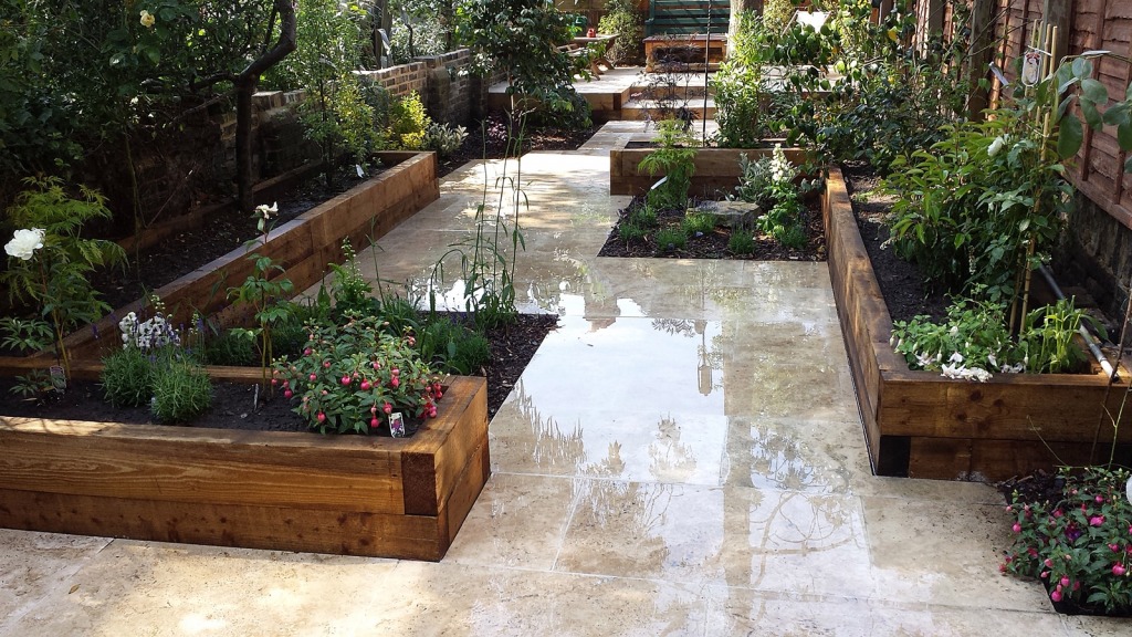 A fantasy garden with polished stone pathways bordered by wooden raised beds. The beds are filled with various green plants, flowers, and shrubs. Trees and additional greenery decorate the background, creating a lush and serene atmosphere.