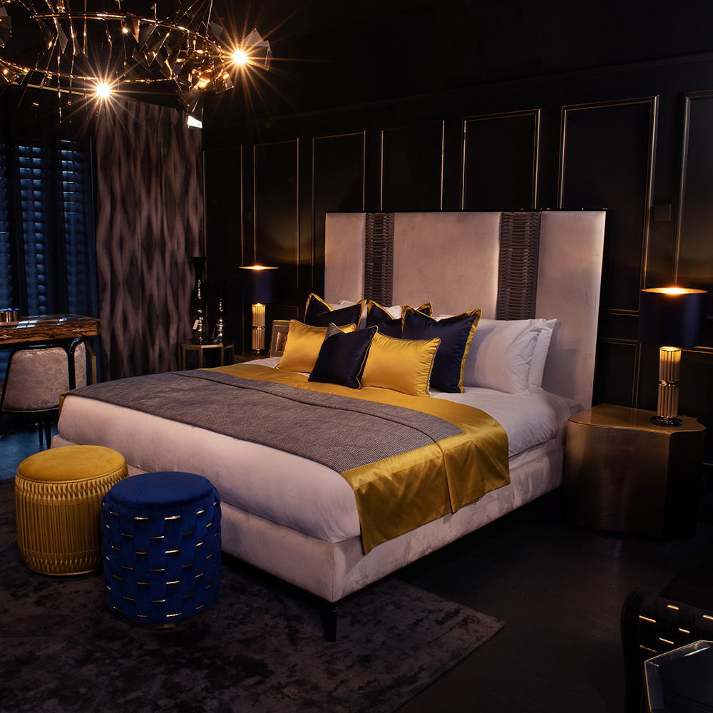 A luxurious bedroom features a large, contemporary bed with white sheets, gray and gold bedding, and multiple pillows. The room is adorned with elegant lighting fixtures, black walls with gold trim, a dark patterned carpet, and a mix of blue and gold ottomans.