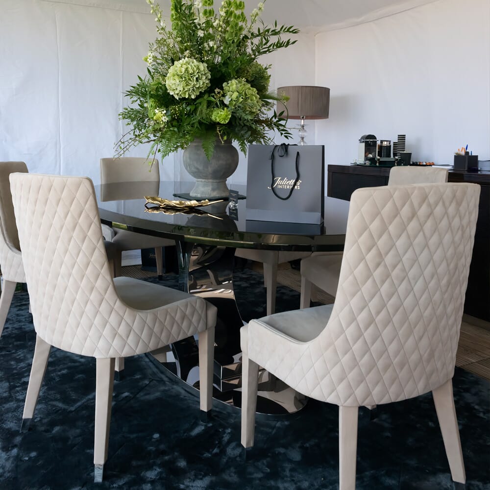 A modern dining room featuring a circular black table surrounded by four beige, quilted chairs. The table displays a large floral arrangement and an elegant shopping bag, showcasing ex display furniture. In the background, there is a sideboard with a coffee machine and decorative items.