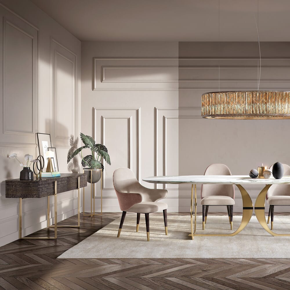 A modern dining room features an elegant, white oval table with gold accents, surrounded by soft pink upholstered chairs with dark legs. A sleek, golden chandelier hangs above. A dark console table and small desks adorned with decor items and potted plants are set against the paneled walls.