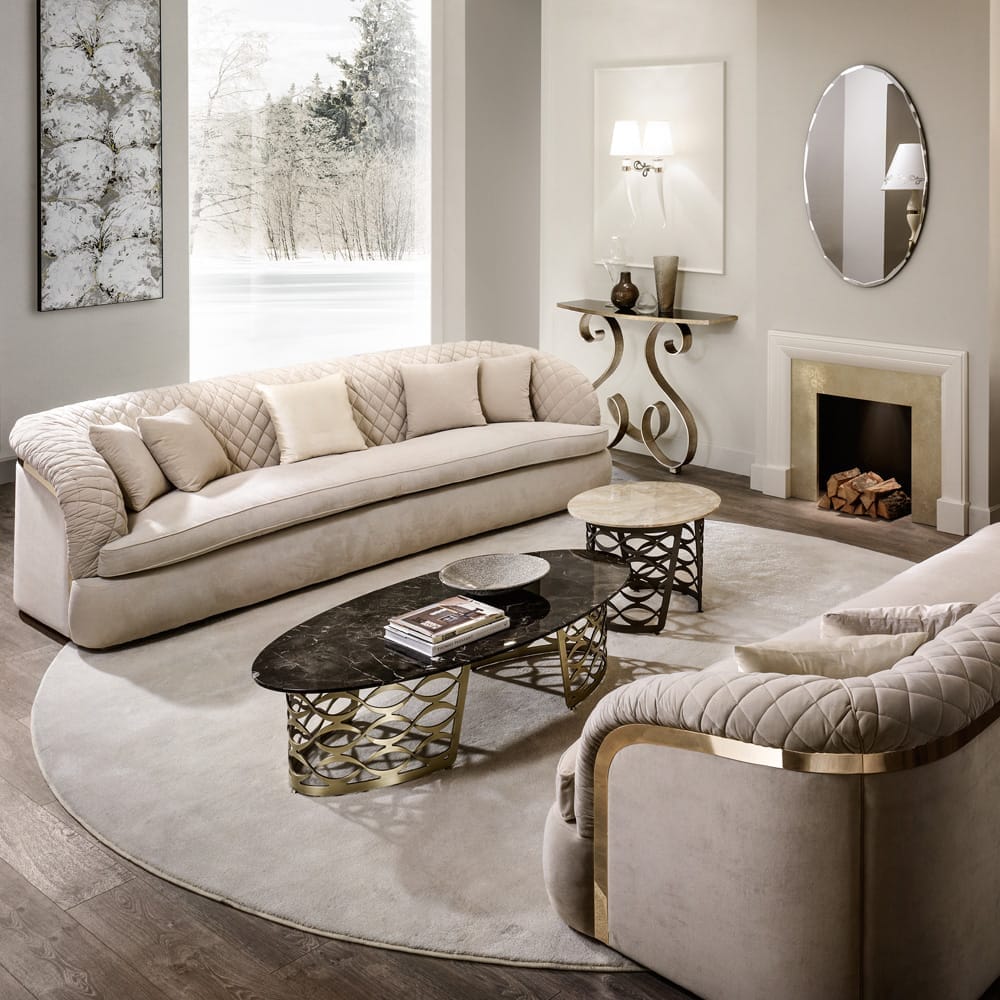 A modern, elegant living room with a cream color palette showcases ex display furniture. It features two plush, beige sofas with quilted backs, a black oval coffee table with a gold geometric base, a gray rug, and a console table with a lamp and mirror. A fireplace with stacked logs completes the cozy ambiance as the large window reveals a snowy outdoor scene.