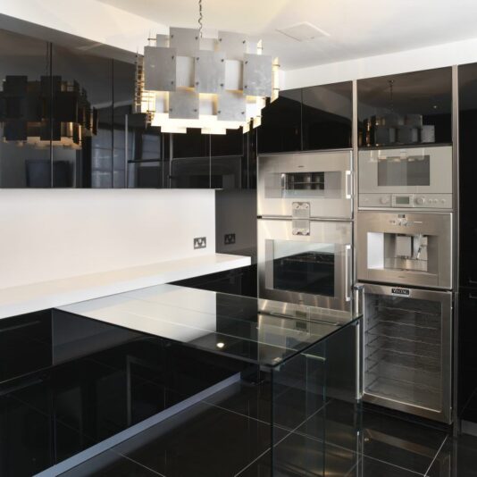 A modern kitchen with sleek black cabinetry and glossy black countertops. It features built-in stainless steel appliances, including an oven, microwave, and coffee machine. A contemporary chandelier with a geometric design hangs from the ceiling.