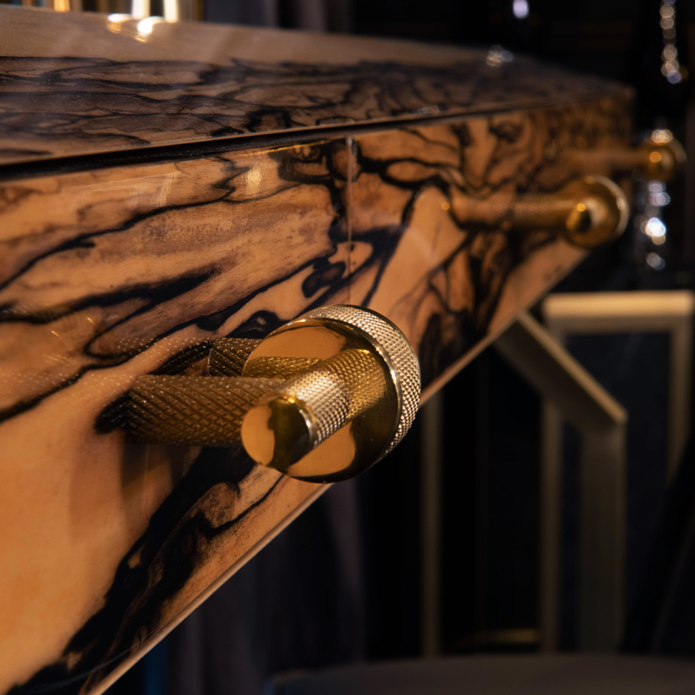 Close-up of a luxurious wooden cabinet from our luxury furniture collection, showcasing intricate grain patterns and a polished, golden metal handle. The cabinet's surface glistens under soft lighting, highlighting its elegant finish and craftsmanship. A hint of a chair is visible in the blurred background.