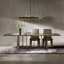 A modern dining room featuring a stylish marble table with a sleek metal base, two chairs with beige cushions, an elegant chandelier with gold accents, and a potted white orchid. The text "Christmas in July" is displayed in the lower-left corner with a small Santa hat, adding a festive touch.