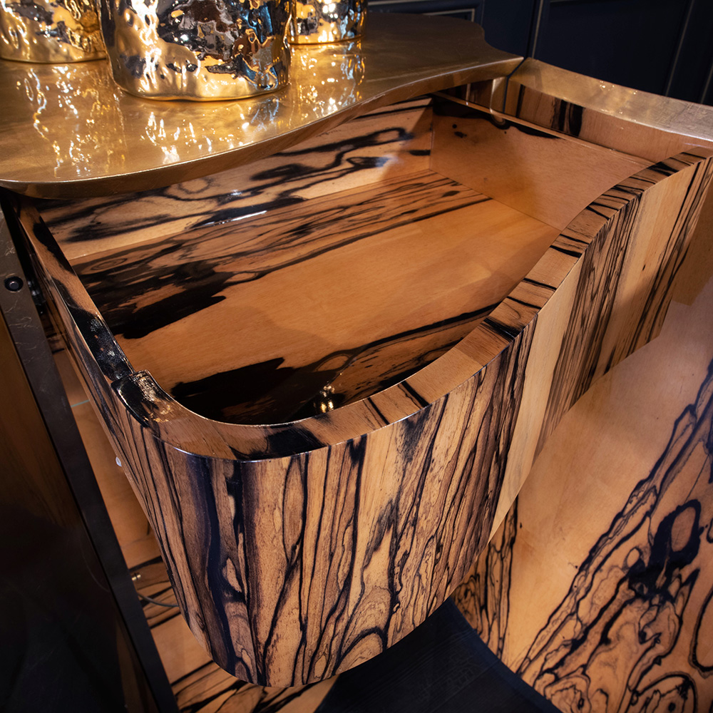 A close-up of a carved wooden drawer made from wood with a striking natural grain featuring black and beige streaks. The top surface has a reflective gold finish, with some gold-colored decorative items placed on it, partially visible in the background—part of our luxury furniture collection.