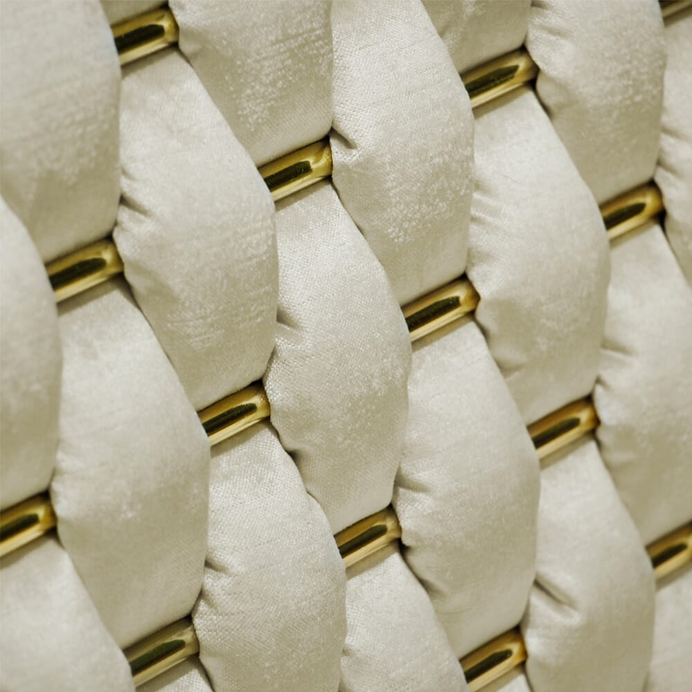 Close-up of a textured surface showing a woven pattern of cushioned, beige fabric strips interlaced with golden metal bars. The pattern features a repetitive, tufted design, creating a soft and luxurious appearance, perfect for any luxury furniture collection.