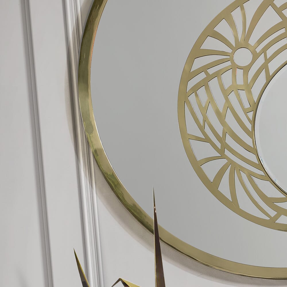 Partial view of a decorative mirror with a gold frame from our luxury furniture collection. The mirror features a circular design with intricate geometric cutouts and a sunburst pattern. The background includes a white paneled wall and part of a gold decor element with pointed accents.