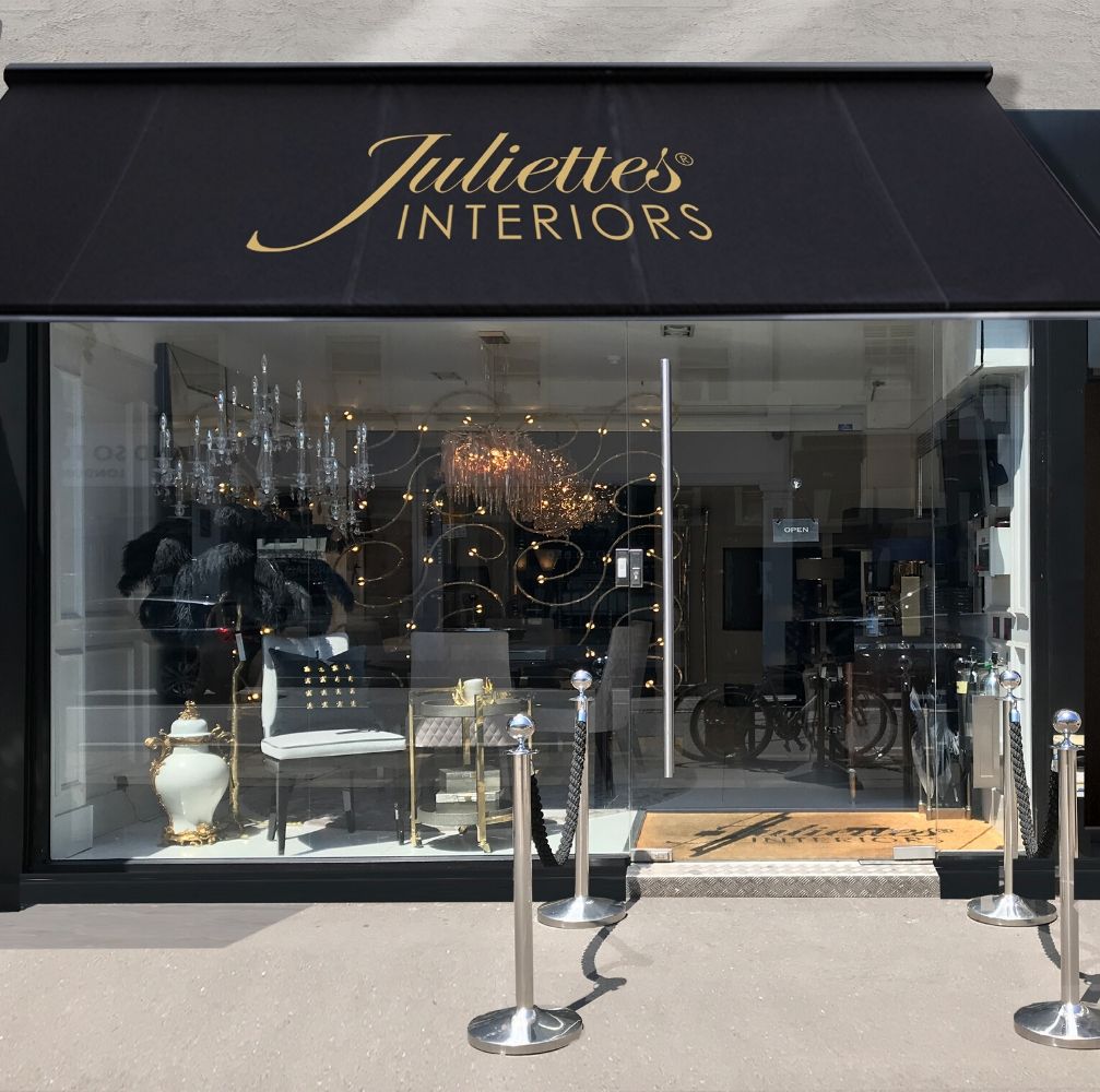 Storefront of Juliettes Interiors with a black awning, gold lettering, and large glass windows displaying luxurious furniture. The entrance is framed by silver stanchions with ropes and a welcome mat. Reflections in the window show street scenes and a bicycle. Offering online consultation for personalized interior design.