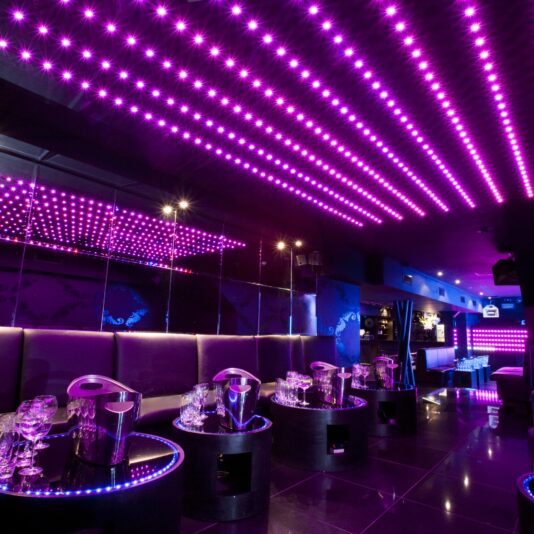 A modern nightclub with purple LED lights on the ceiling. The room is furnished with black cushioned seats, round tables with ice buckets and glasses, and mirrored walls reflecting the colorful lighting. The ambiance is vibrant and futuristic.