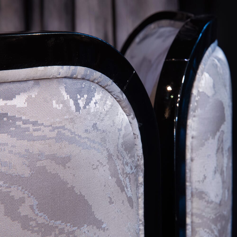 Close-up of an upholstered chair from the luxury furniture collection, featuring a curved, glossy black frame. The fabric has a light gray, abstract pattern resembling marbled texture. The background is softly blurred, drawing attention to the chair's design and texture details.